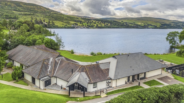 A lakeside building with a reception area, surrounded by lush greenery and a large body of water. - The Kenmore Club (Aberfeldy)