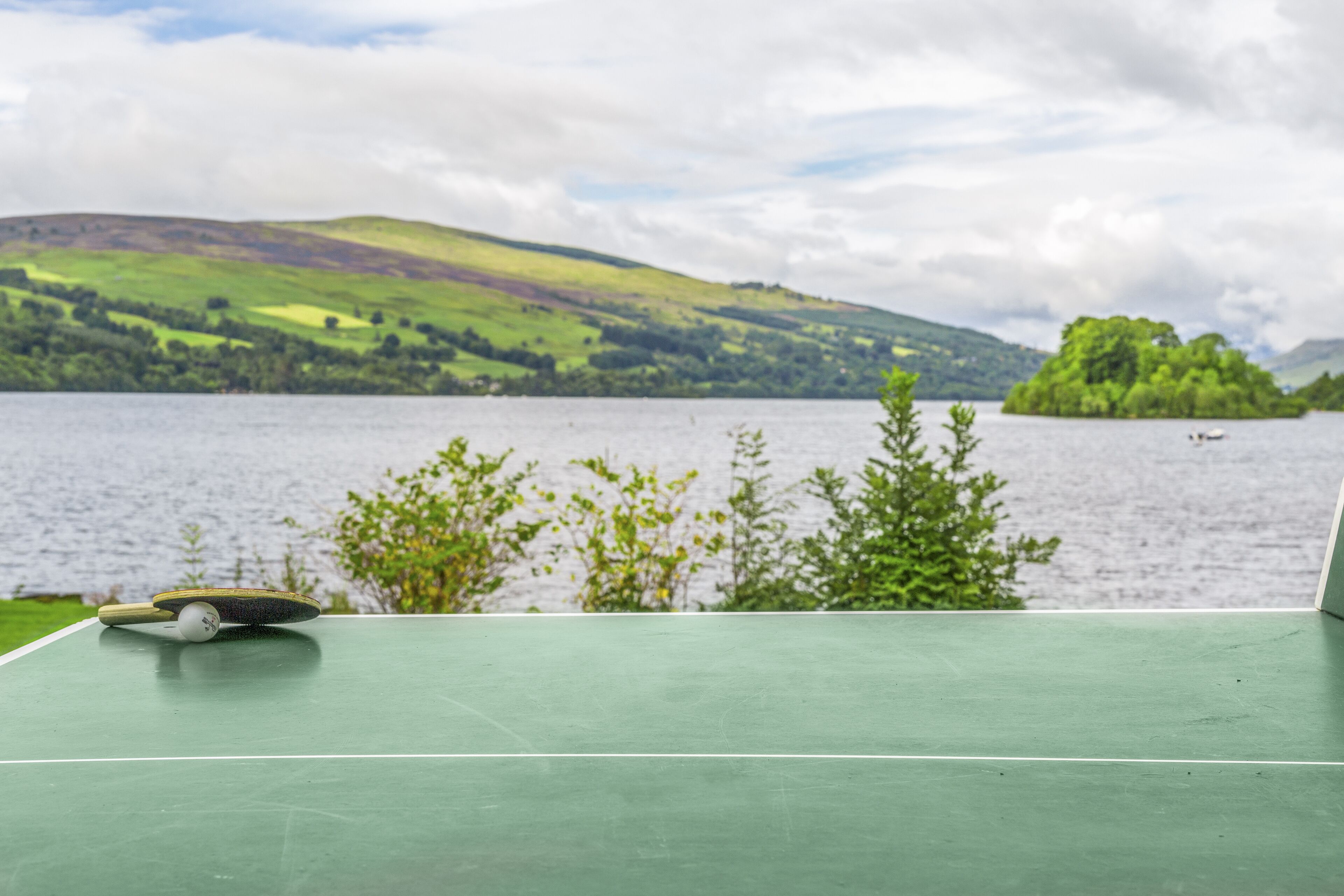 A green tennis court with a net, overlooking a lake with hills and a boat in the distance.