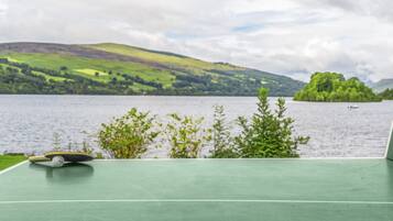 A green tennis court with a net, overlooking a lake with hills and a boat in the distance.