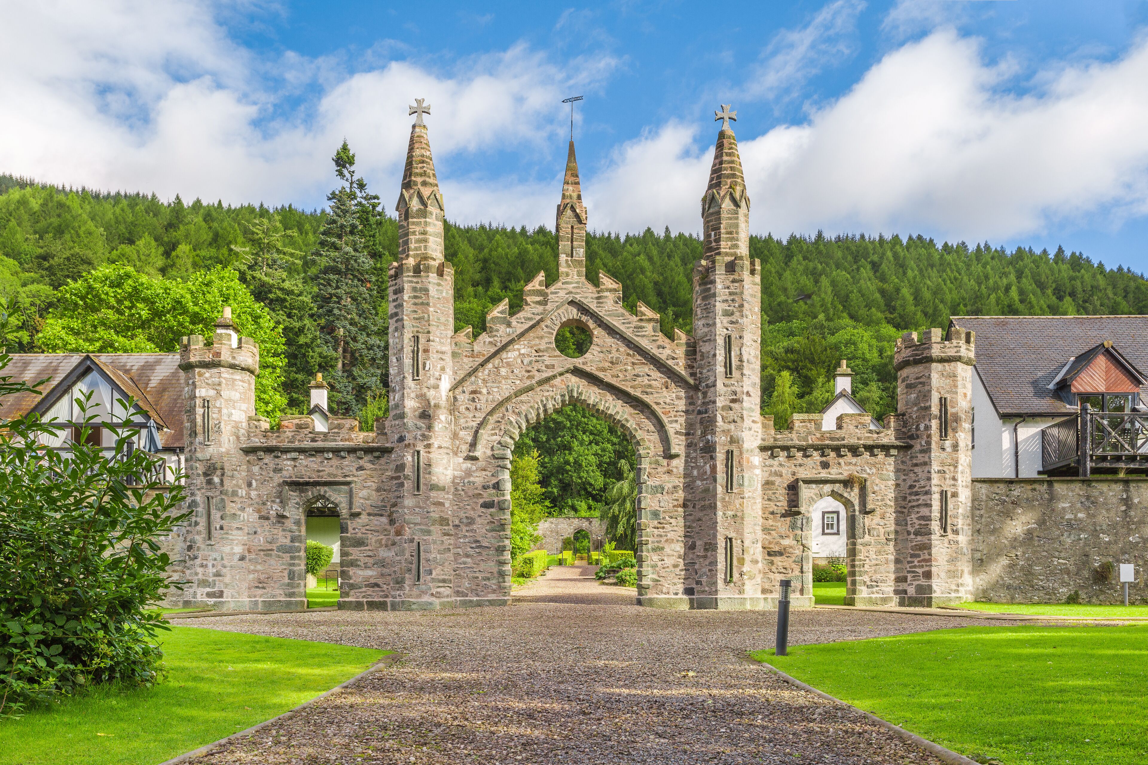 A stone church with a central archway and three spires, surrounded by a well-maintained lawn and a forested hill in the background.