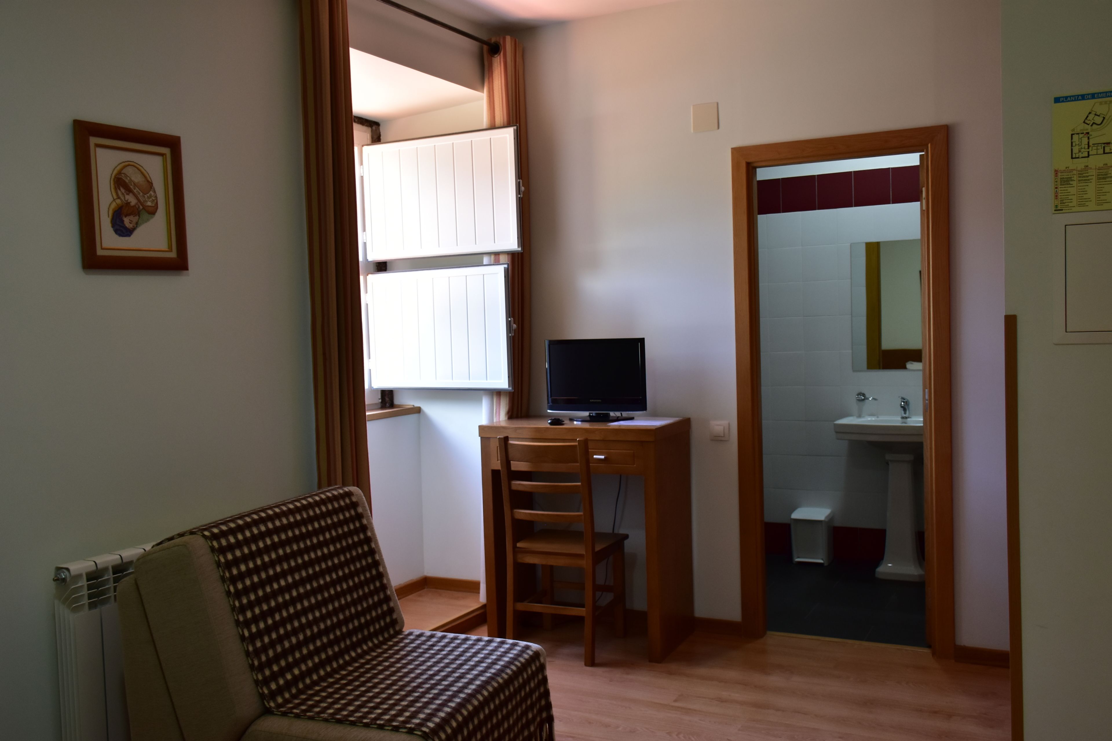 A small living room with a sofa, a wooden desk with a computer, a window with curtains, and a door leading to a bathroom.