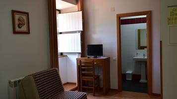 A small living room with a sofa, a wooden desk with a computer, a window with curtains, and a door leading to a bathroom.