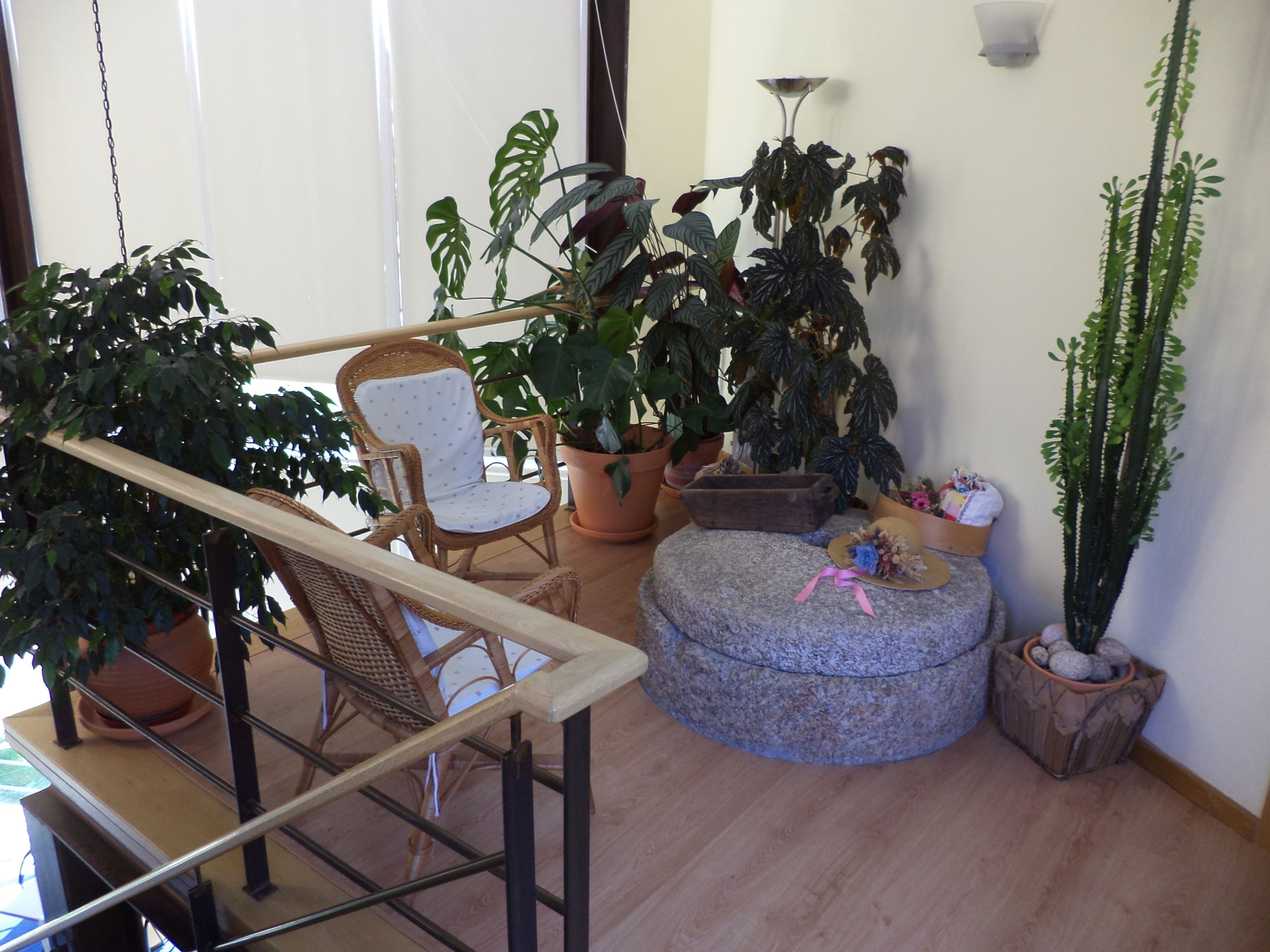 Un balcón con una silla de mimbre, plantas en macetas y un otomano de piedra.