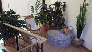 Un balcón con una silla de mimbre, plantas en macetas y un otomano de piedra.