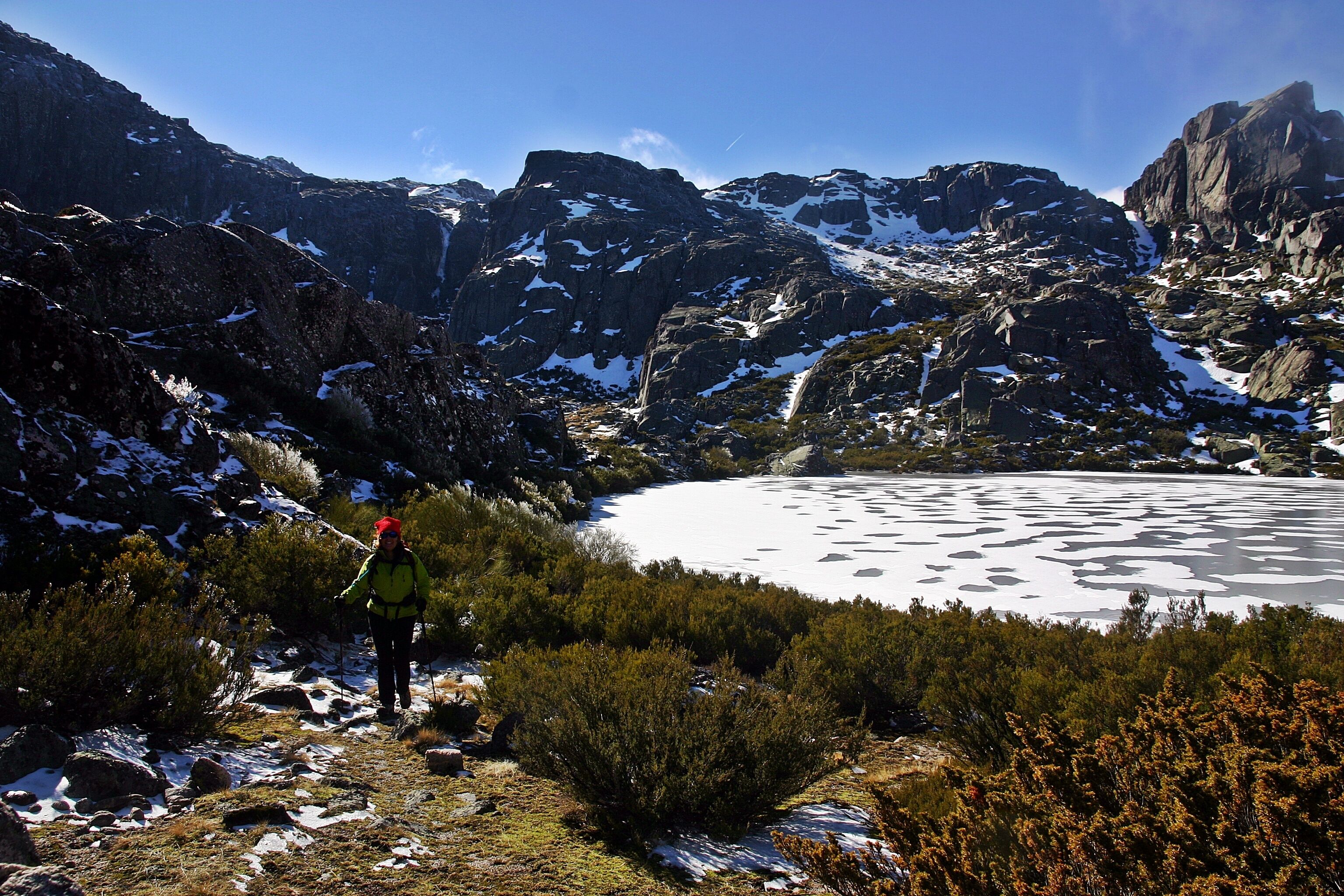 Ein Wanderer auf einem felsigen Pfad mit einem schneebedeckten See und Bergen im Hintergrund.