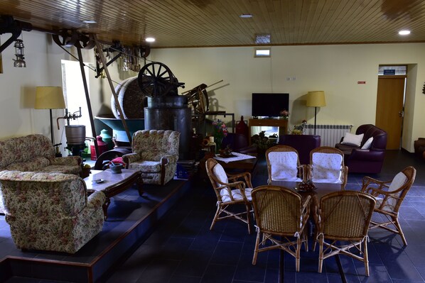 A living room with floral armchairs, a dining table with wicker chairs, a large wooden wheel, and a television.