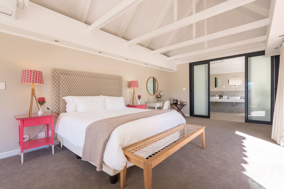 Un dormitorio moderno con una cama grande, mesitas de noche, un banco, un escritorio y un baño visible a través de una puerta de cristal.