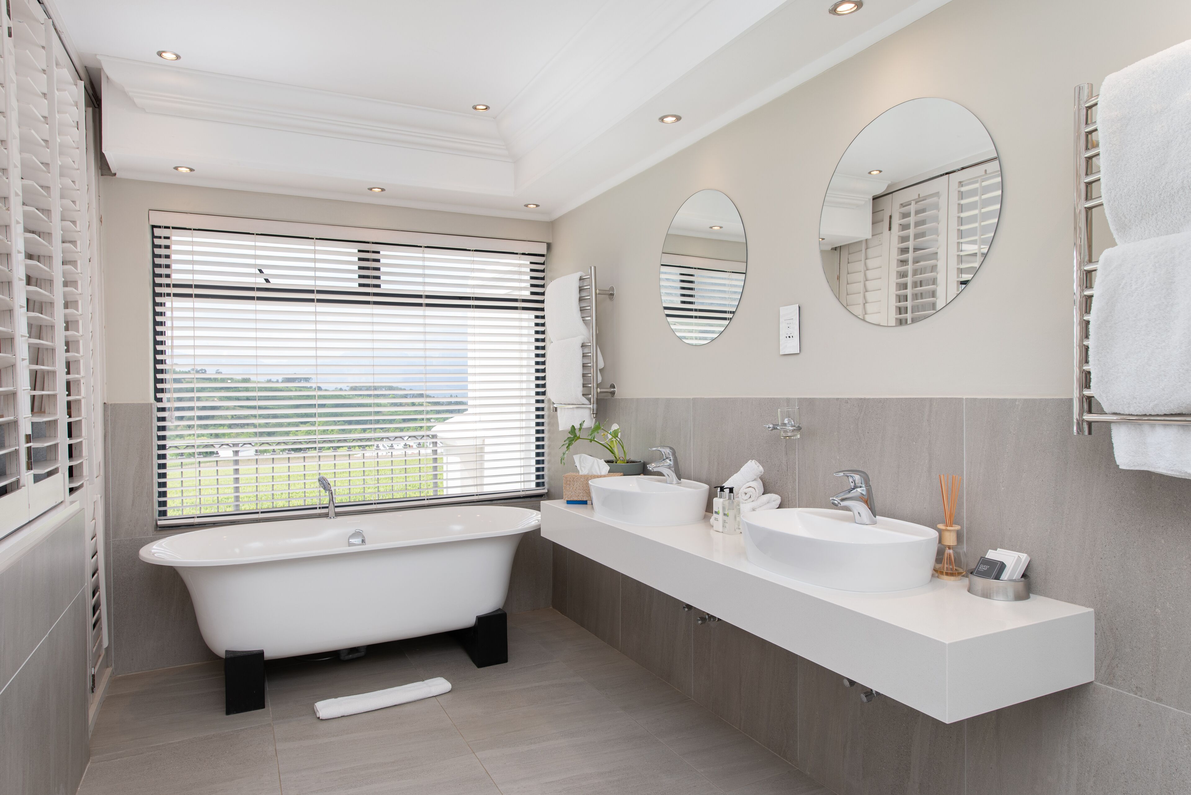 Ein modernes Badezimmer mit einer freistehenden Badewanne, zwei Waschbecken und einem großen Fenster mit Jalousien.