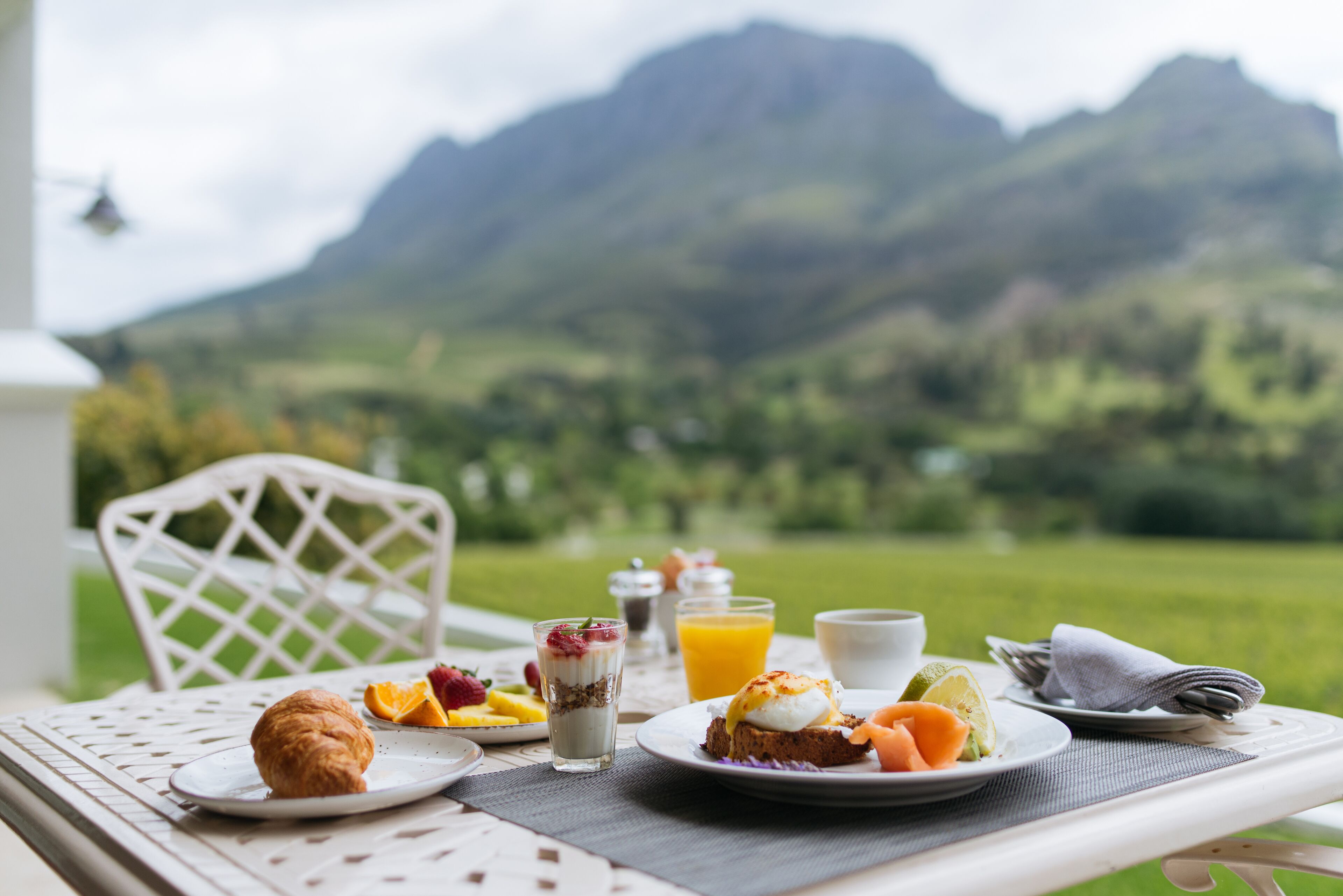 Een ontbijttafel met croissants, fruit, yoghurt parfait en sinaasappelsap, met een prachtig berglandschap op de achtergrond.
