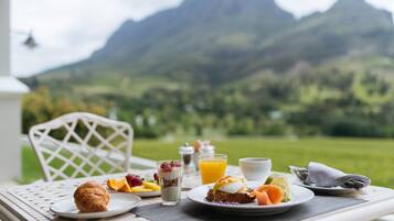 Een ontbijttafel met croissants, fruit, yoghurt parfait en sinaasappelsap, met een prachtig berglandschap op de achtergrond.