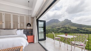 Quarto com uma cama grande, mesa de cabeceira e vista para uma paisagem montanhosa através de uma porta de vidro.