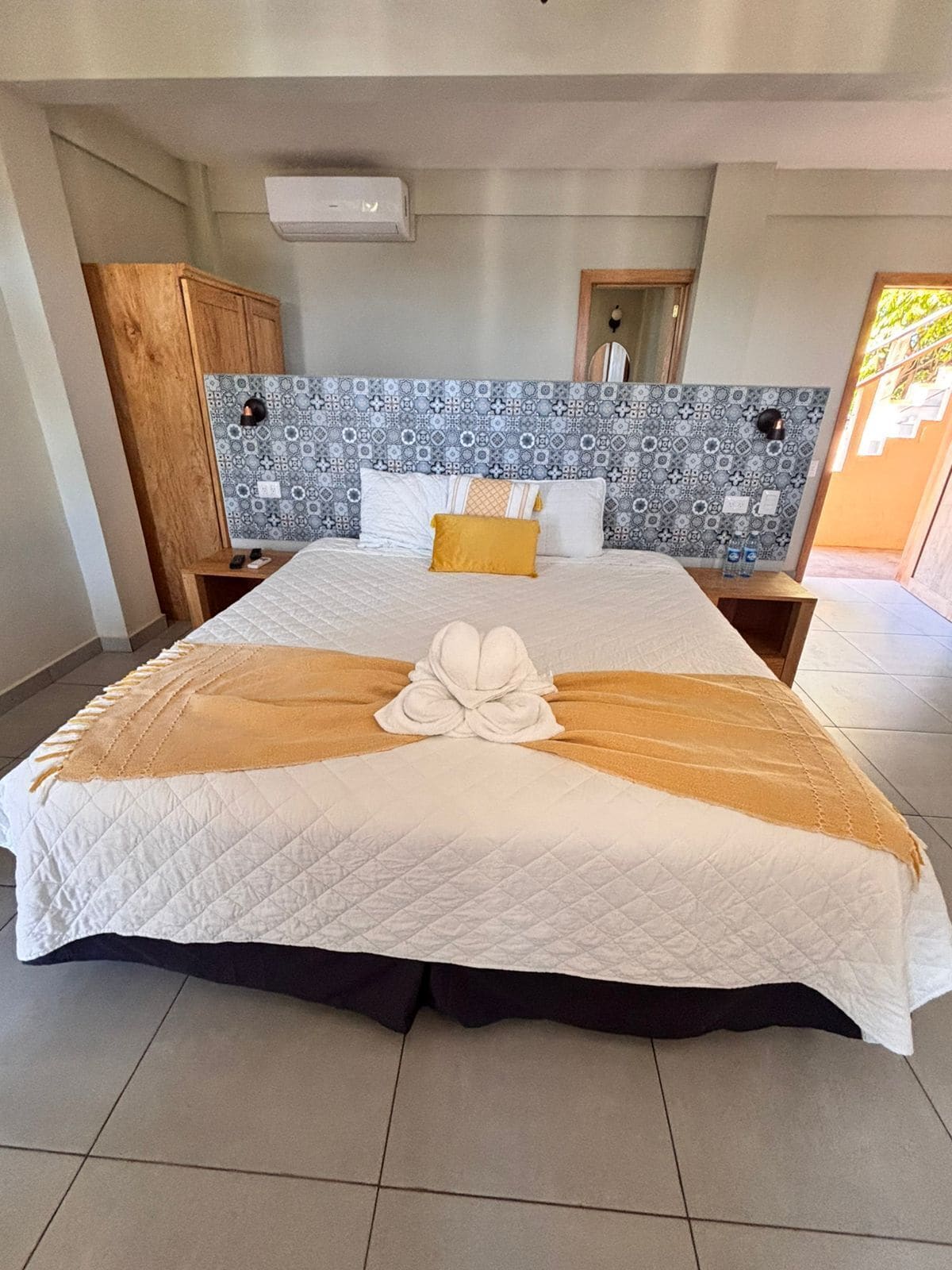 A hotel room with a large bed, a wooden wardrobe, a patterned headboard, and a tiled floor.