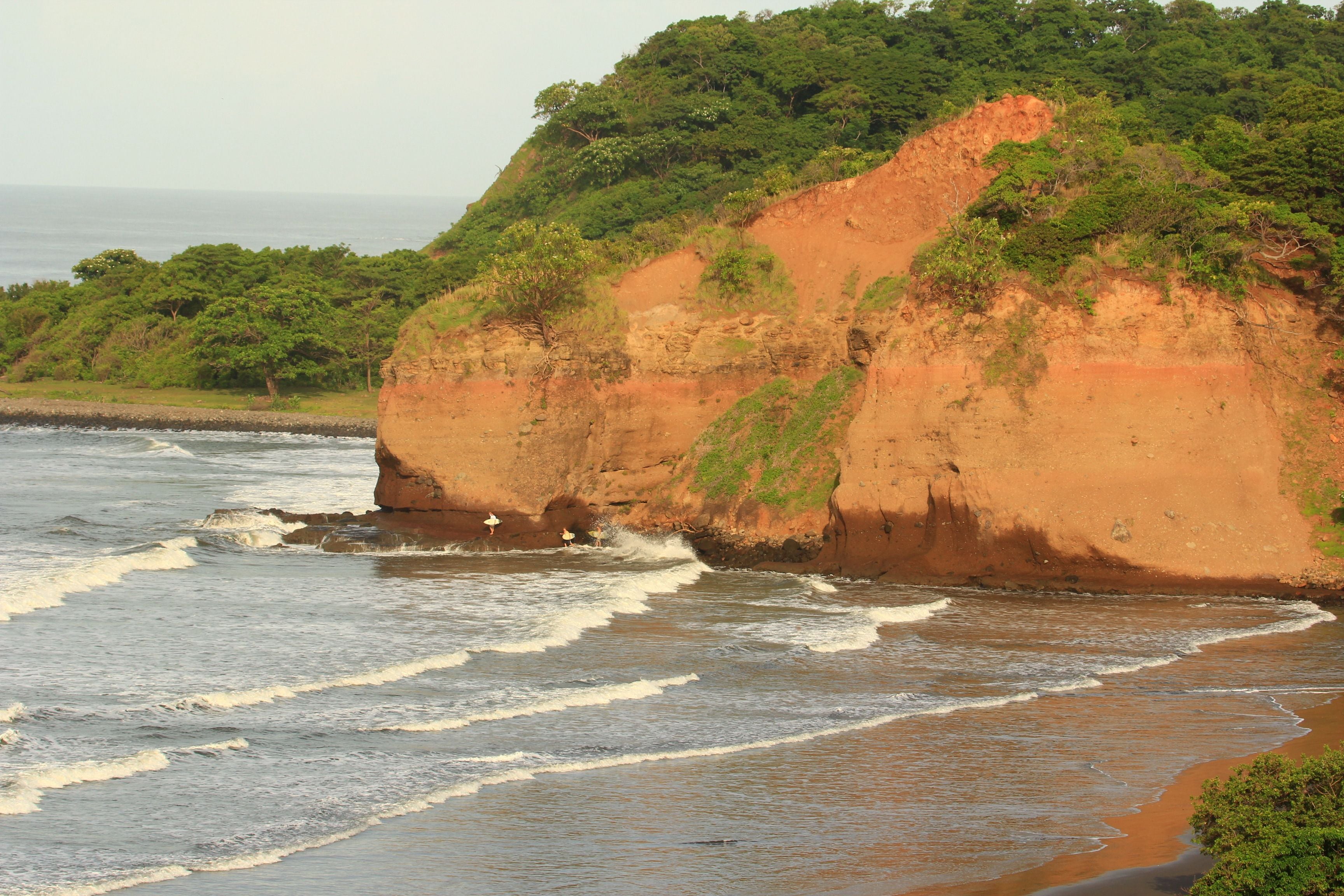 Un paisaje costero con una playa arenosa, olas y un acantilado con vegetación.