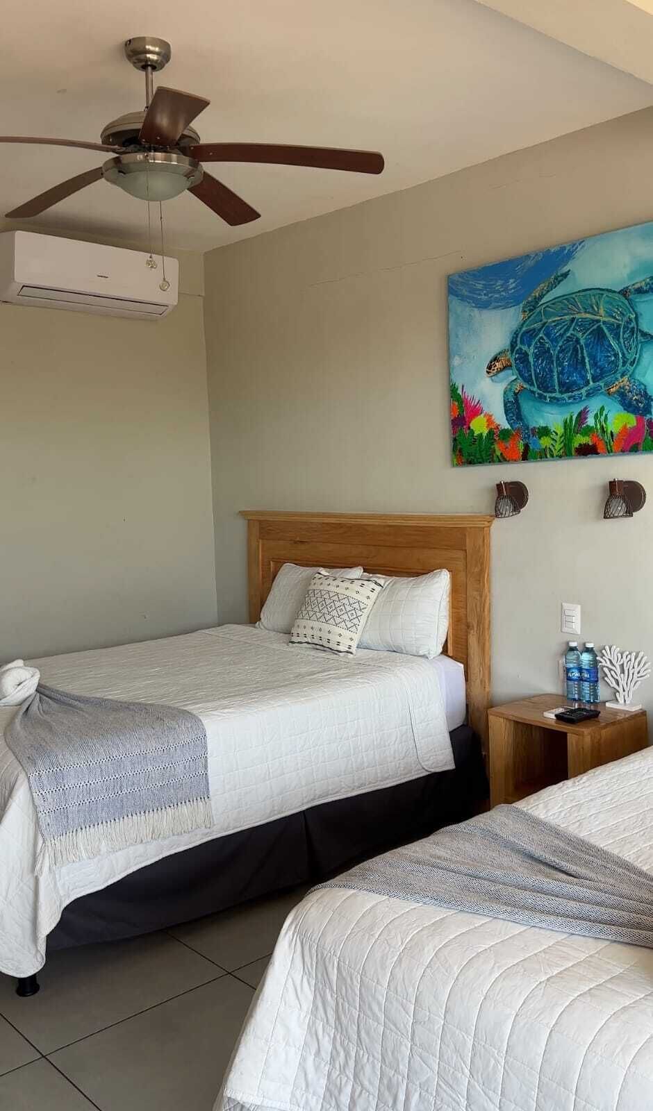 Habitación de hotel con dos camas, ventilador de techo, equipo de aire acondicionado, un cuadro de una tortuga y una mesita de noche de madera con una botella y un teléfono.