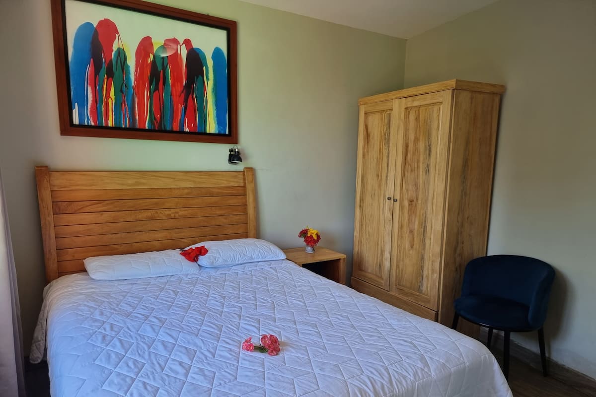 Uma cama bem arrumada com um edredom branco acolchoado, cabeceira de madeira, guarda-roupa de madeira e uma pequena mesa de cabeceira com um vaso de flores.