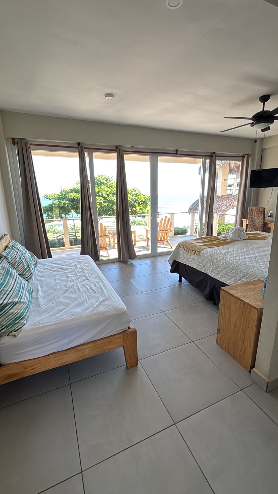 A hotel room with two beds, a balcony with a view of the sea, and a ceiling fan.