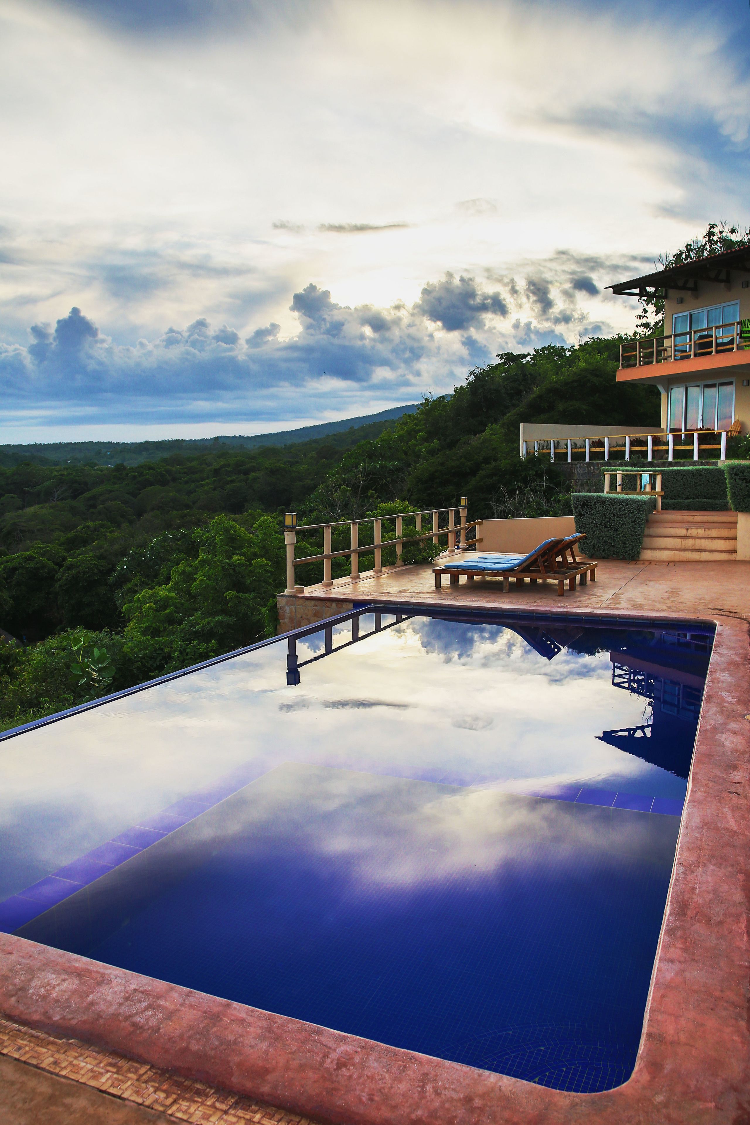 A swimming pool with a clear view of the surrounding landscape, a lounge chair, and a staircase leading to a balcony.