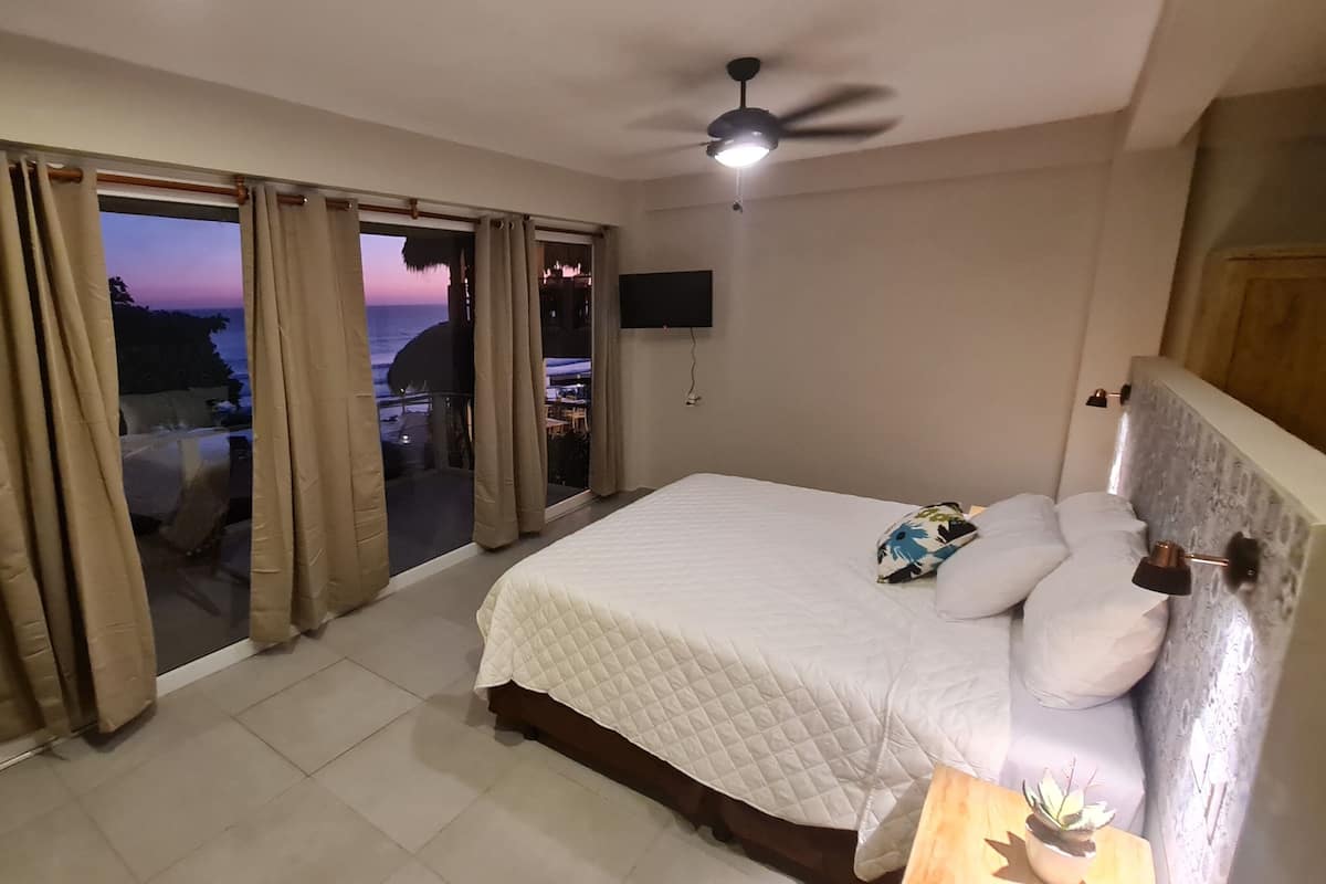 Quarto com cama, ventilador de teto, TV fixada na parede e vista para o exterior.