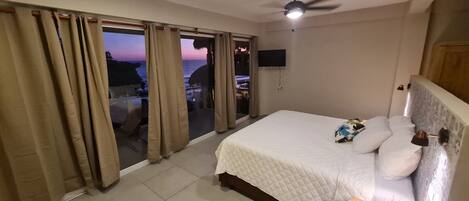 A bedroom with a bed, a ceiling fan, a TV mounted on the wall, and a view of the outside.