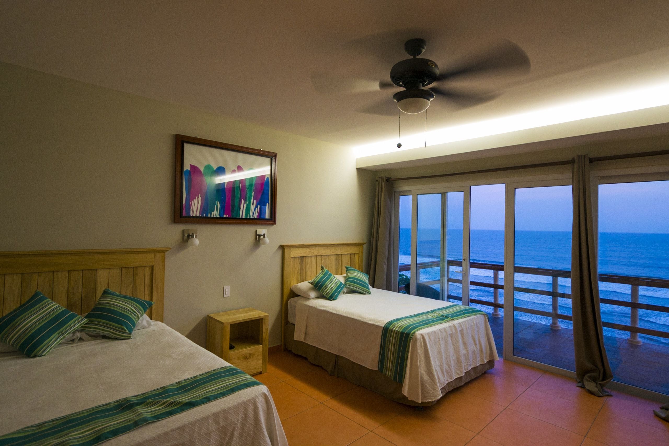 A hotel room with two beds, a ceiling fan, a painting, and a balcony with a view of the ocean.