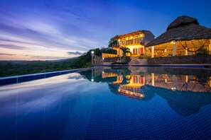 Un resort lujoso con una amplia piscina infinita que refleja el edificio y construcciones con techo de paja.