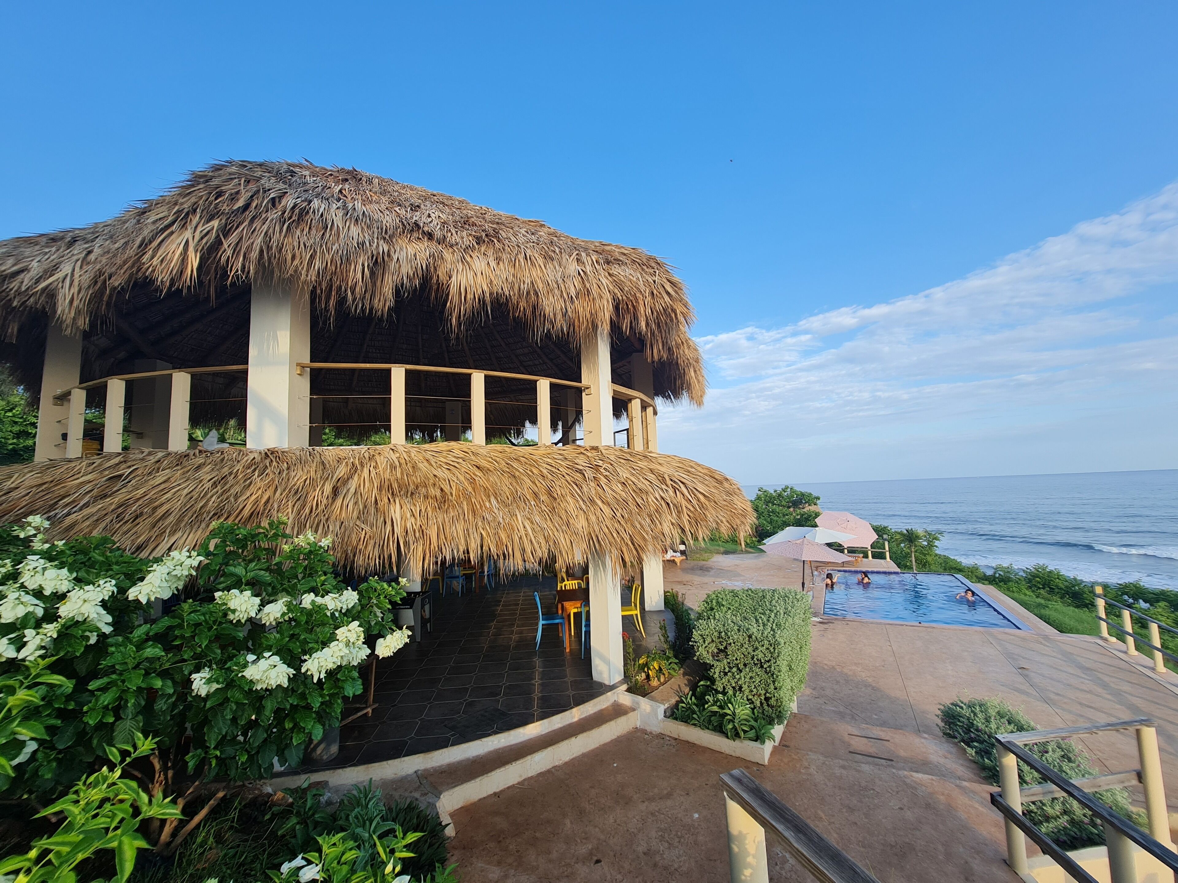 A resort with a thatched-roof building, a swimming pool, and a view of the ocean.