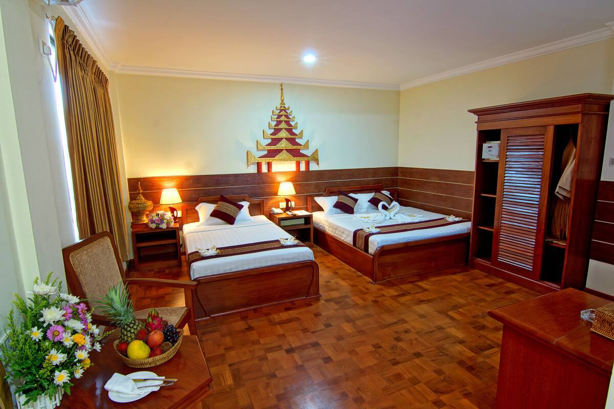 A hotel room with two beds, wooden flooring, a traditional decorative wall piece, a wardrobe, and a small table with a fruit basket and flowers.