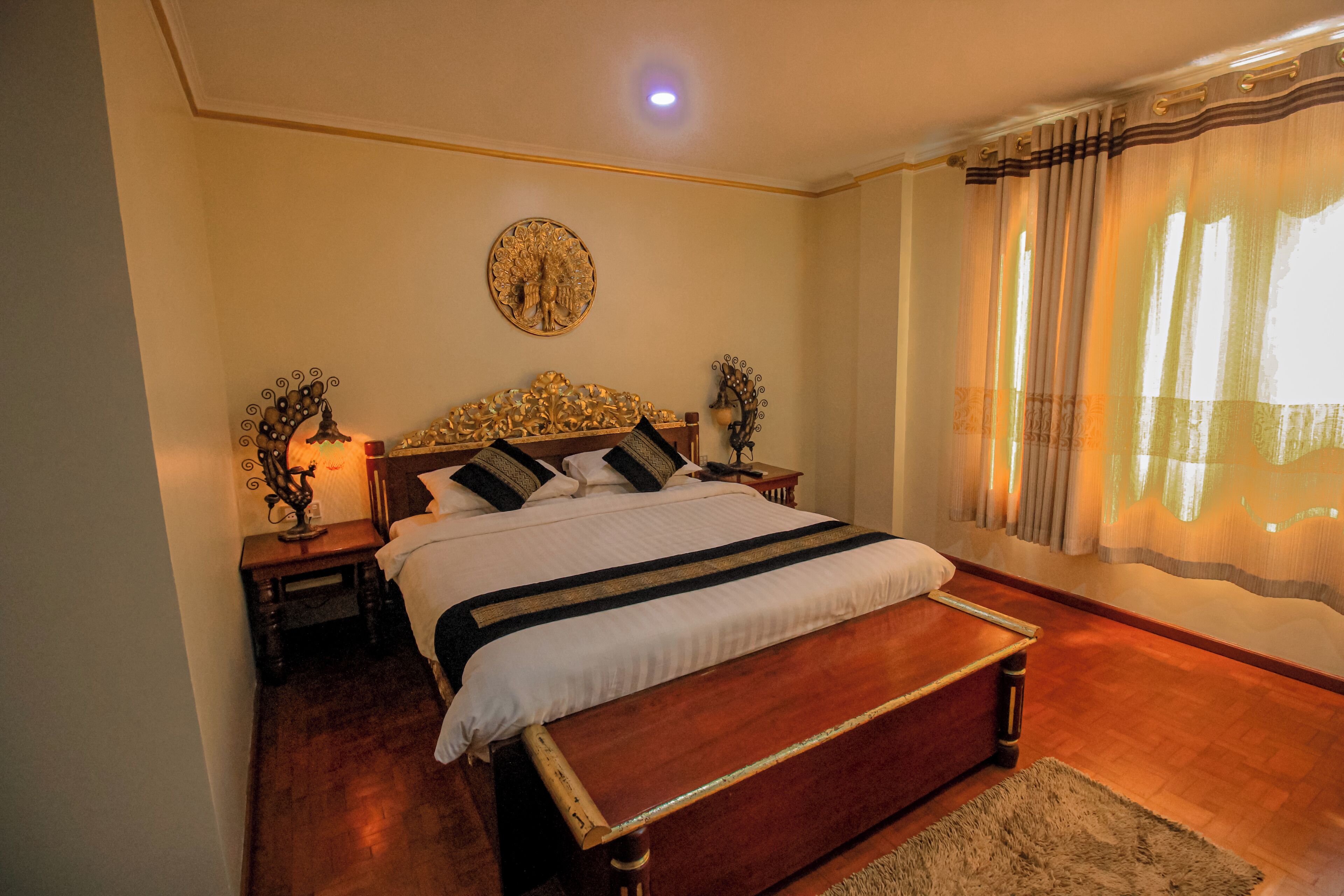 Un dormitorio con una cama grande, cabecera de madera, dos mesitas de noche con lámparas, un adorno circular en la pared y una ventana con cortinas.