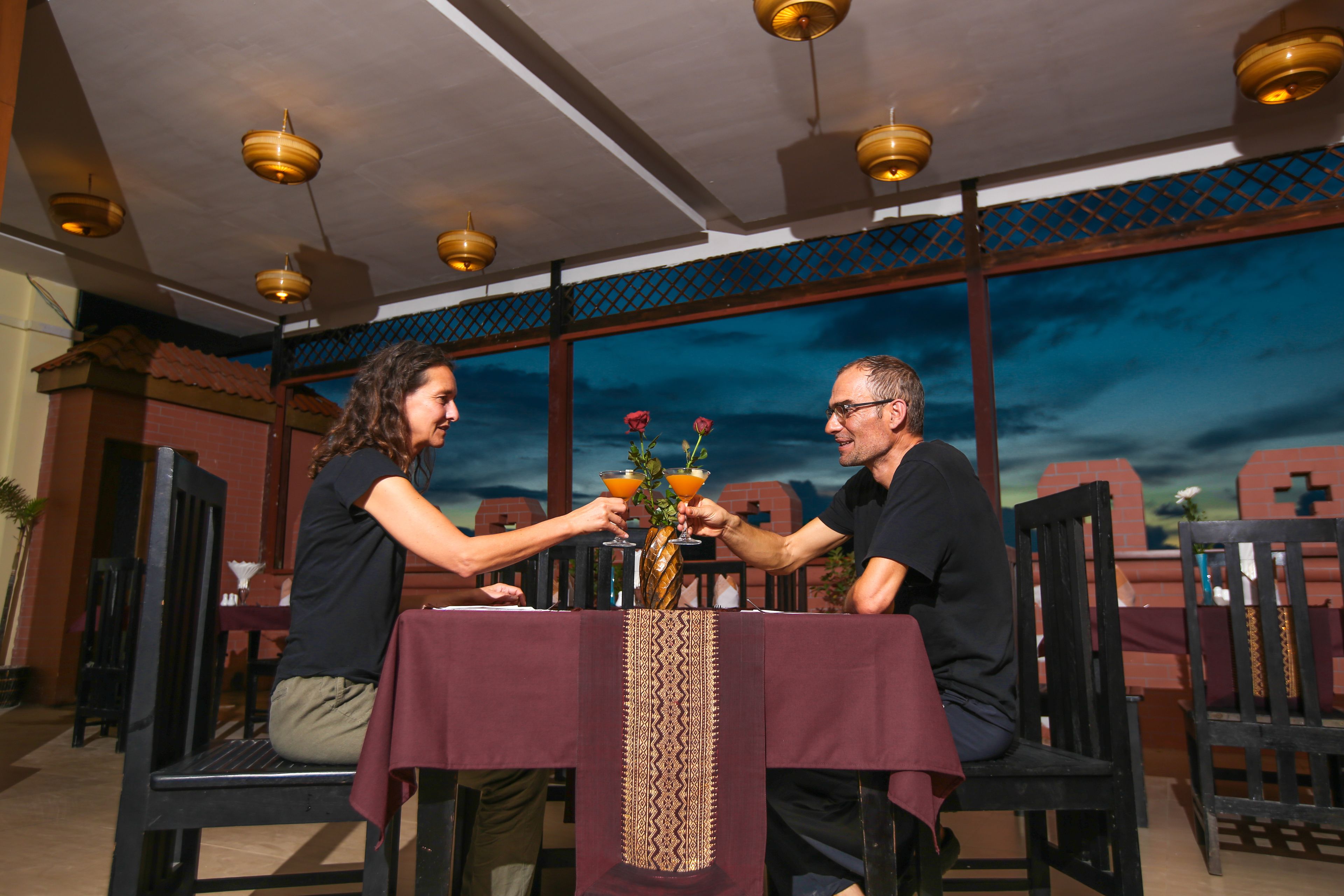 Deux personnes sont assises à une table recouverte d’une nappe à motifs. Un vase rempli de fleurs est posé sur la table, et de grandes fenêtres offrent une vue sur le coucher de soleil.