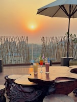 Vista del atardecer desde una terraza con una mesa que sostiene tres bebidas y un paraguas grande.