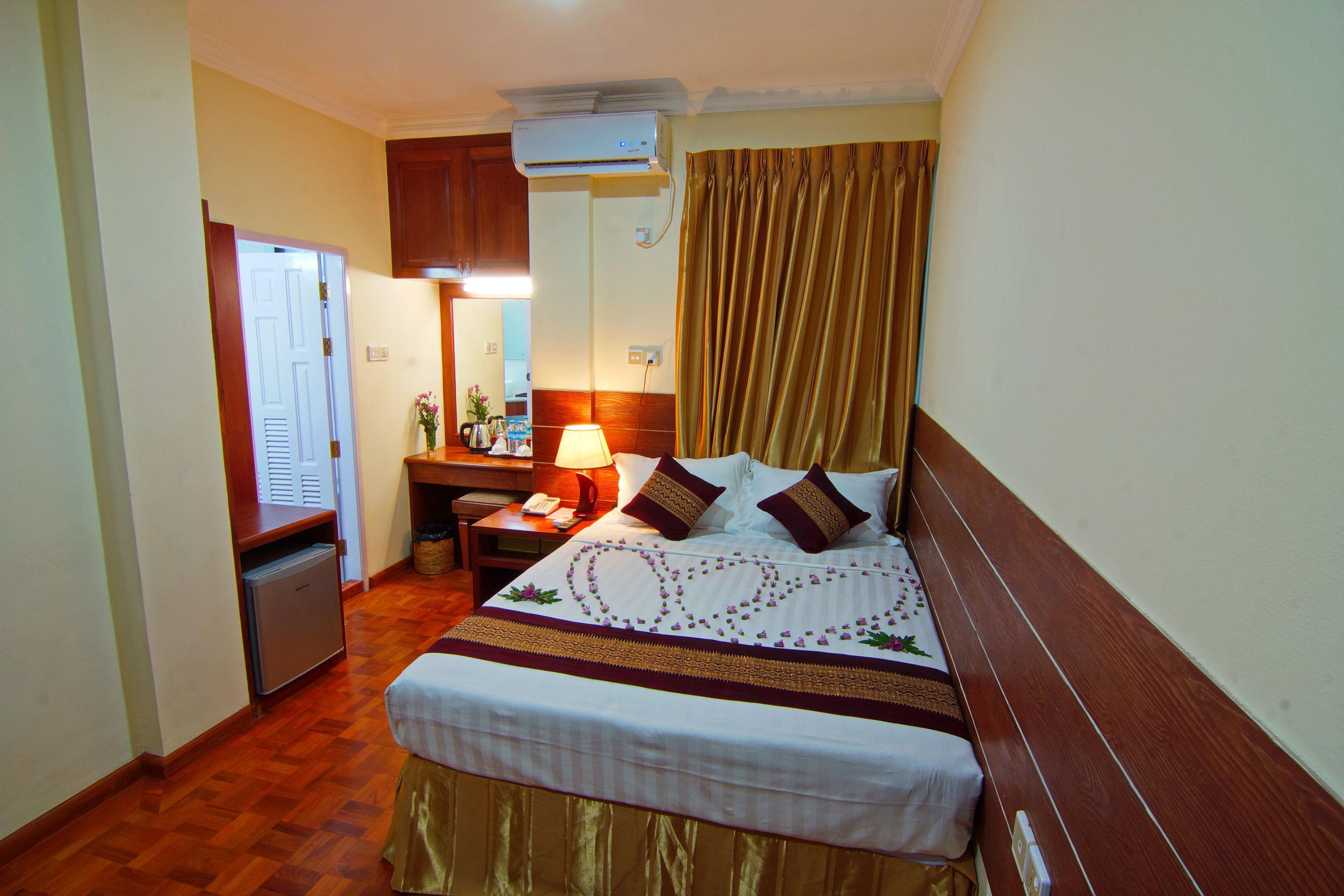 A hotel room with a bed, a desk, a lamp, a mirror, and an air conditioning unit.