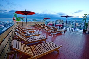 Terraza en la azotea con sillones de madera y sombrillas rojas, con vistas a un paisaje urbano.