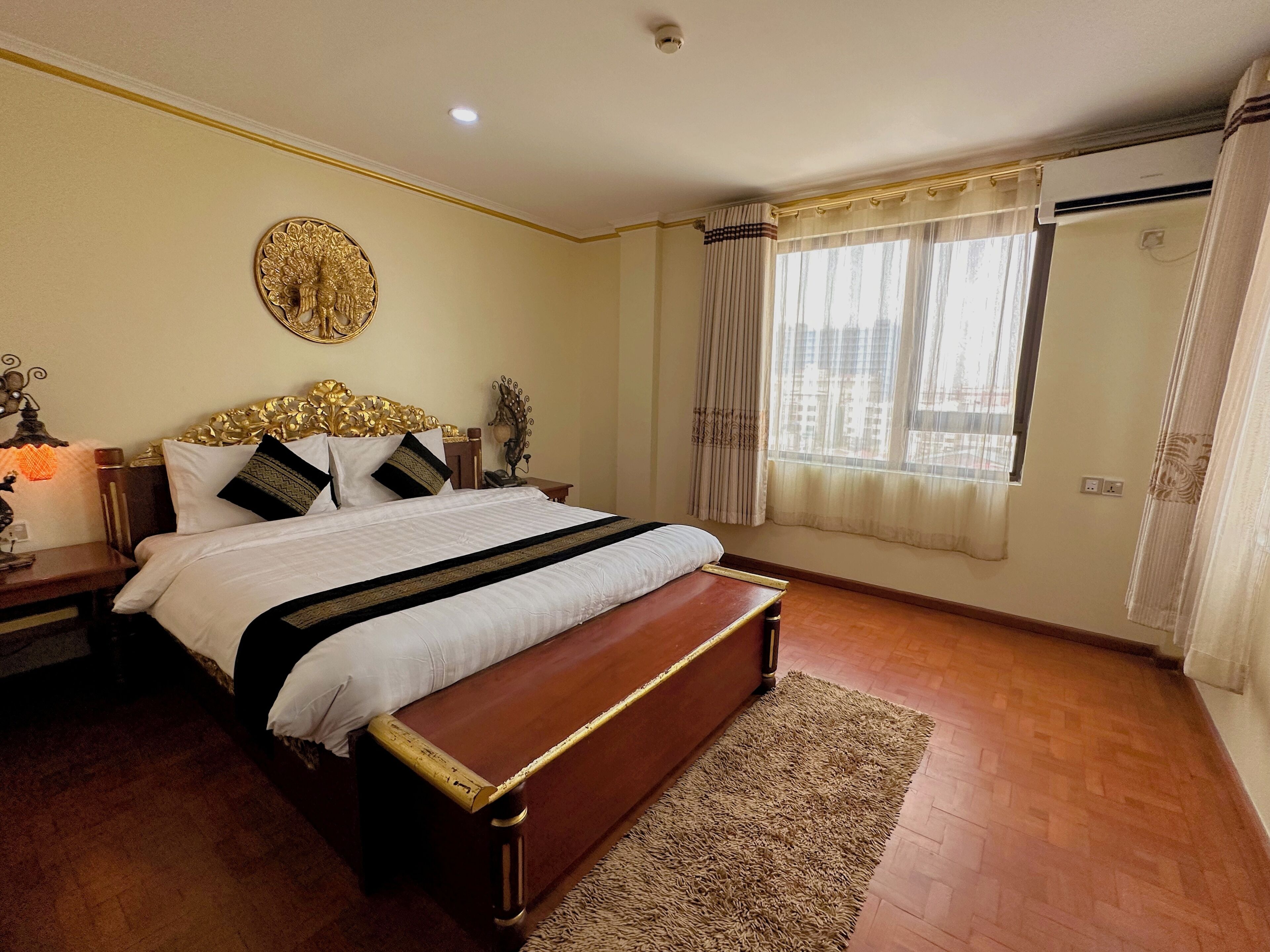 A bedroom with a large bed, a bench, a decorative wall piece, and a window with curtains.