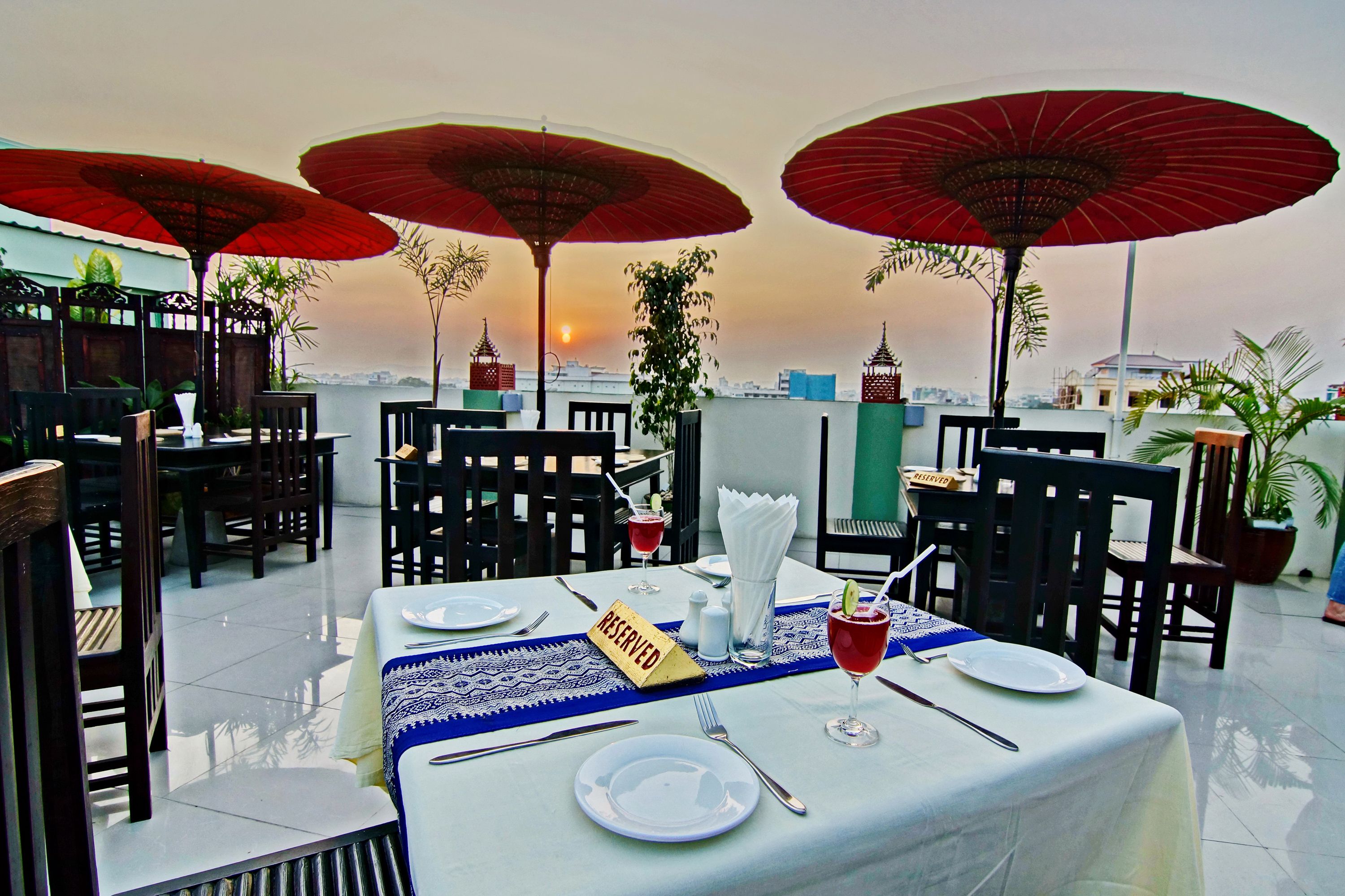 A rooftop restaurant with tables set for dining, red umbrellas, and a view of the sunset.