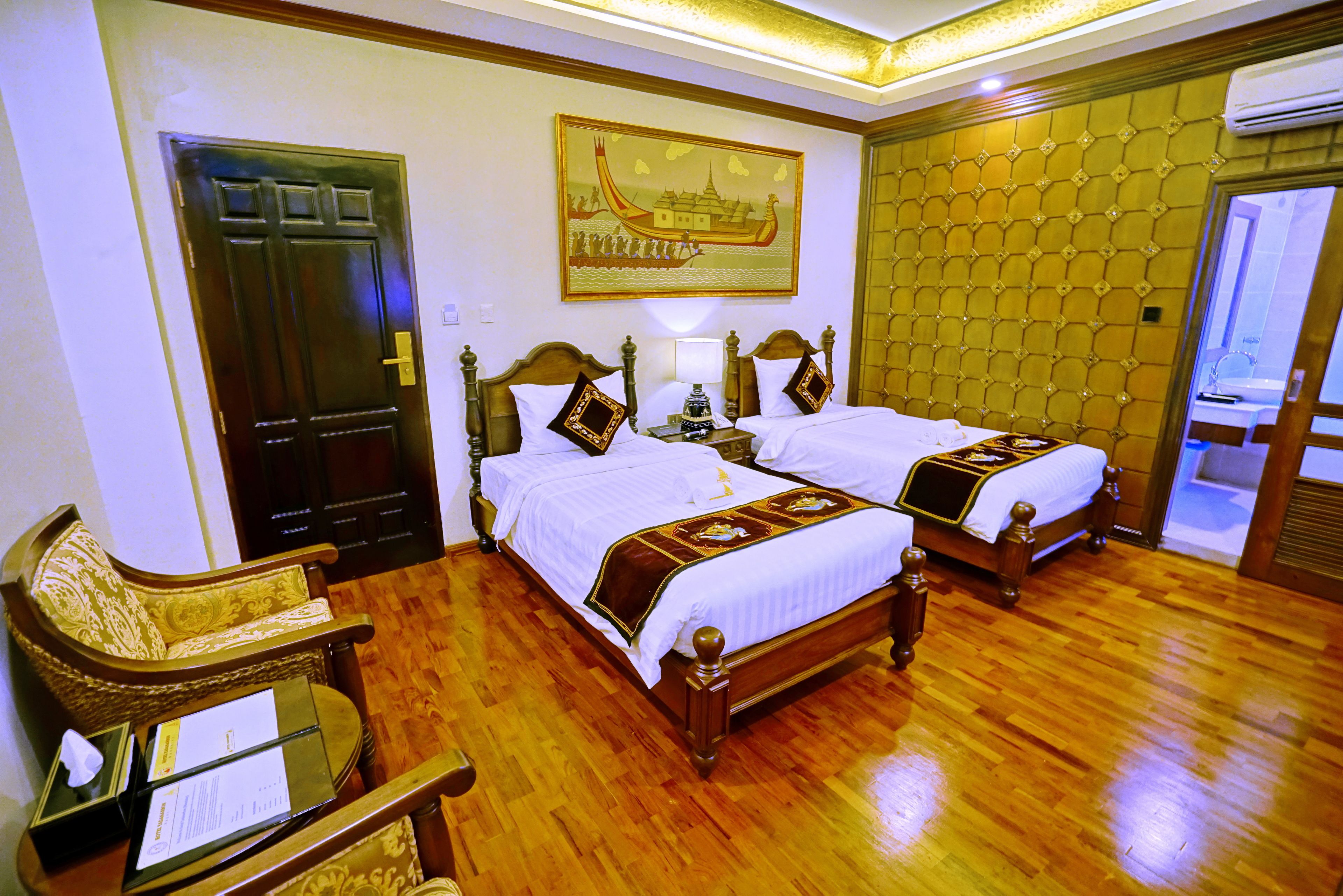 A hotel room with two wooden beds, a desk with a chair, a door, and a wall-mounted air conditioning unit.