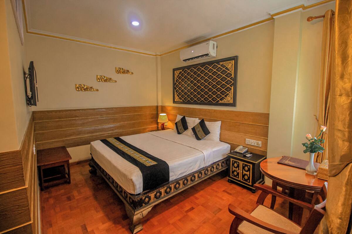 A hotel room with a large bed, a bedside table, a chair, a small table, and a wall-mounted air conditioner.