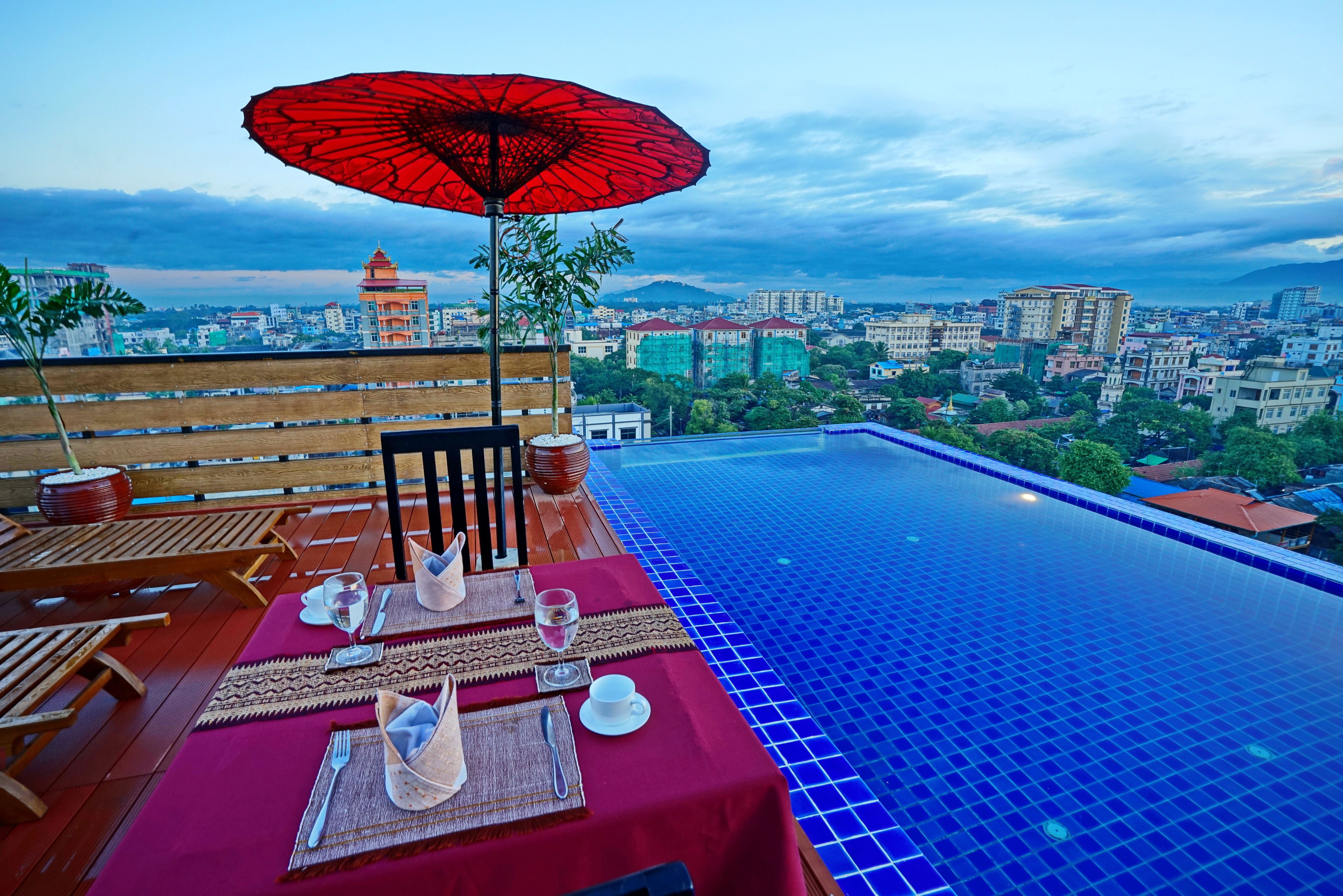 An infinity pool with a city view, a table set for two with a red umbrella, and wooden furniture.