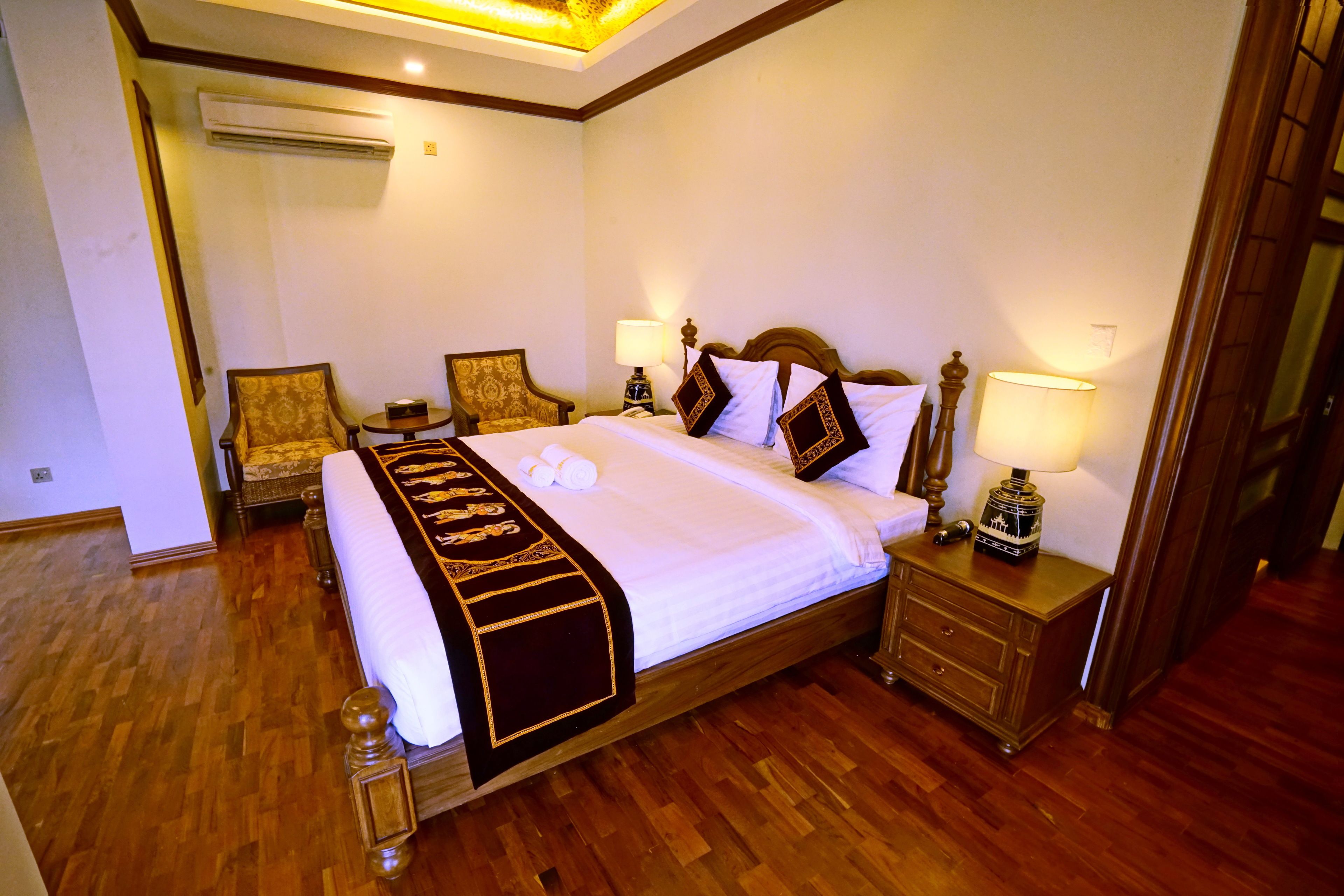 A hotel room with a large bed, wooden headboard, two bedside tables with lamps, a wooden floor, and an air conditioning unit on the wall.