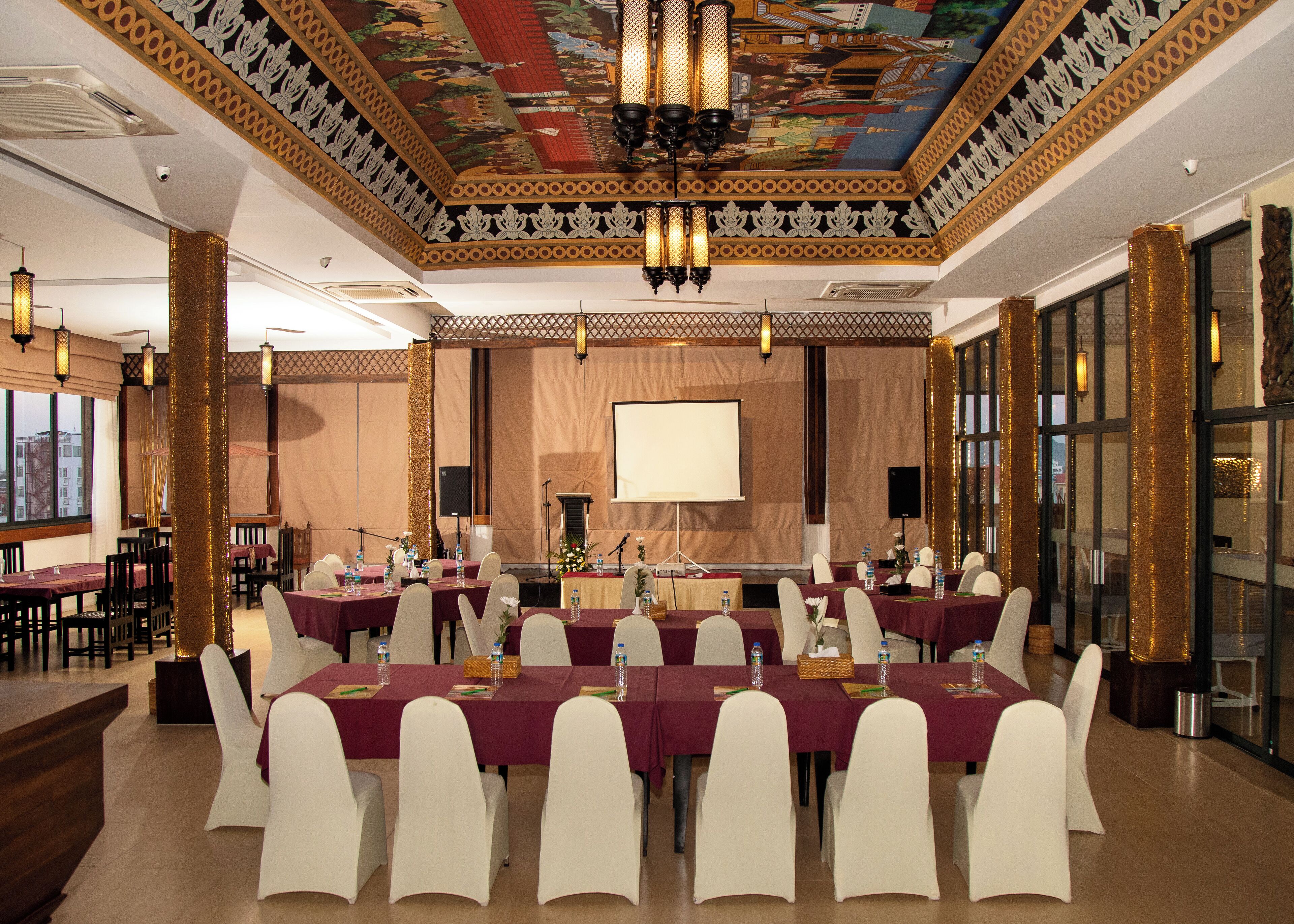 A banquet hall with tables set for a formal event, a stage with a projector screen, and a decorative ceiling.