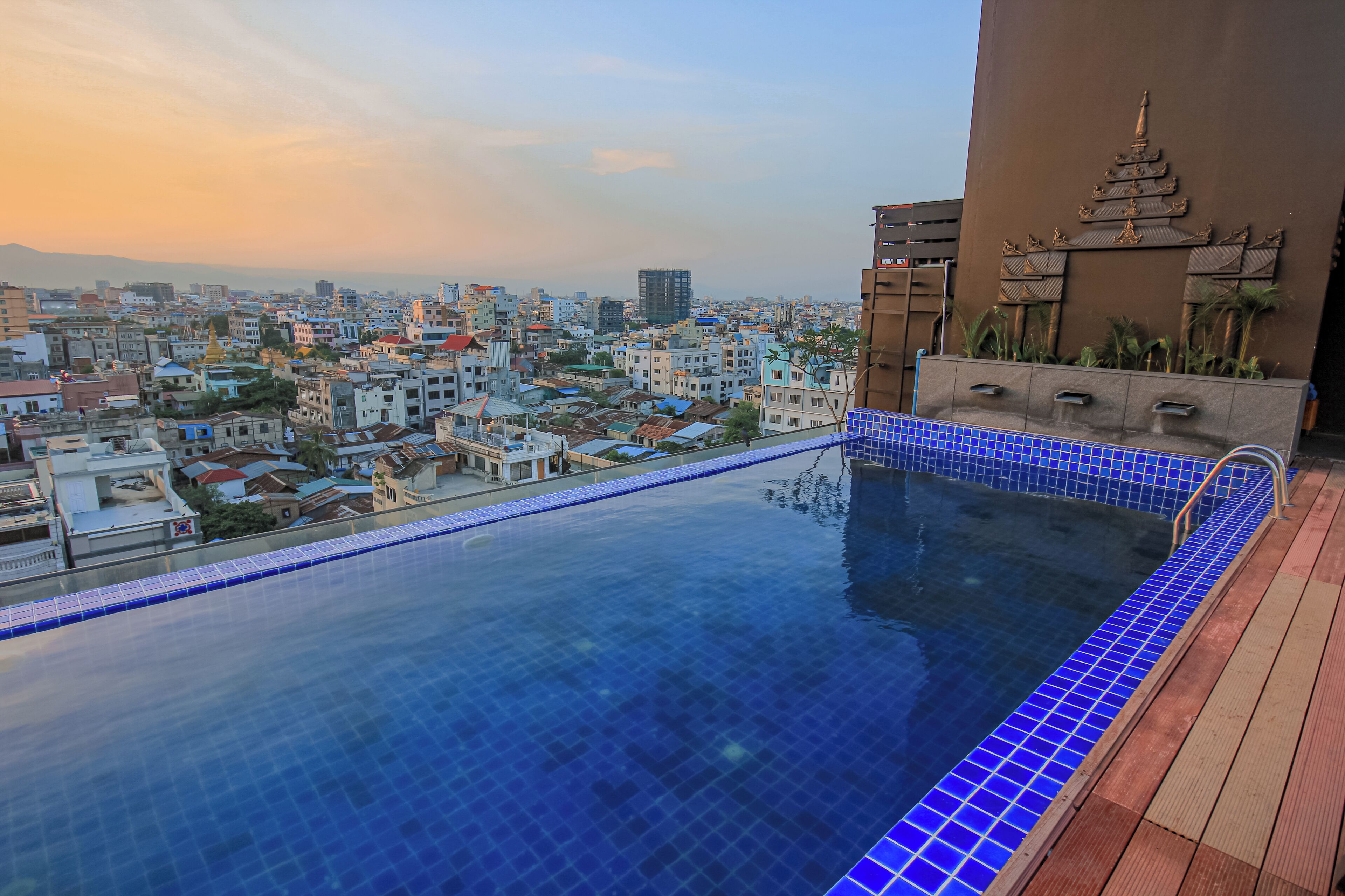 A rooftop infinity pool with a city view, a pagoda-style wall decoration, and a wooden deck.