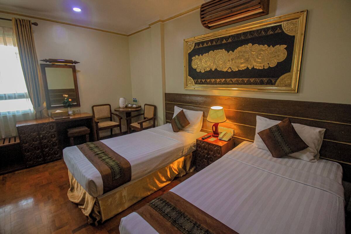 A hotel room with two beds, a desk, a chair, a lamp, and a large decorative wall piece.