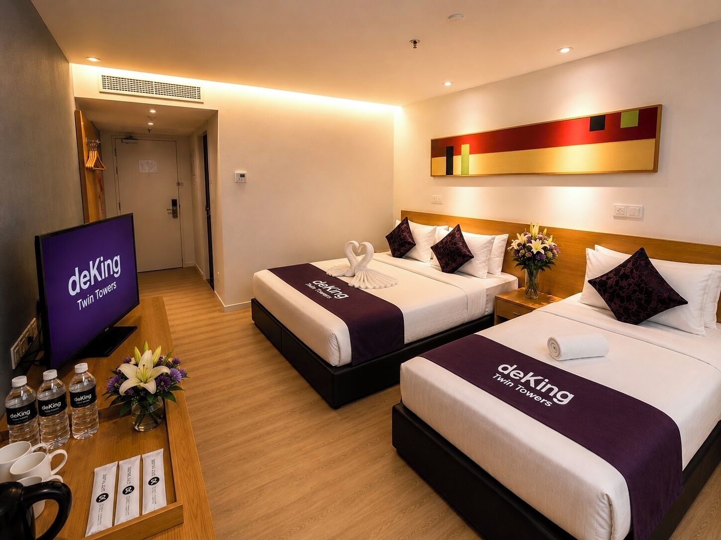A hotel room with two beds, a TV, a desk with water bottles and a vase of flowers, and a wall art piece.
