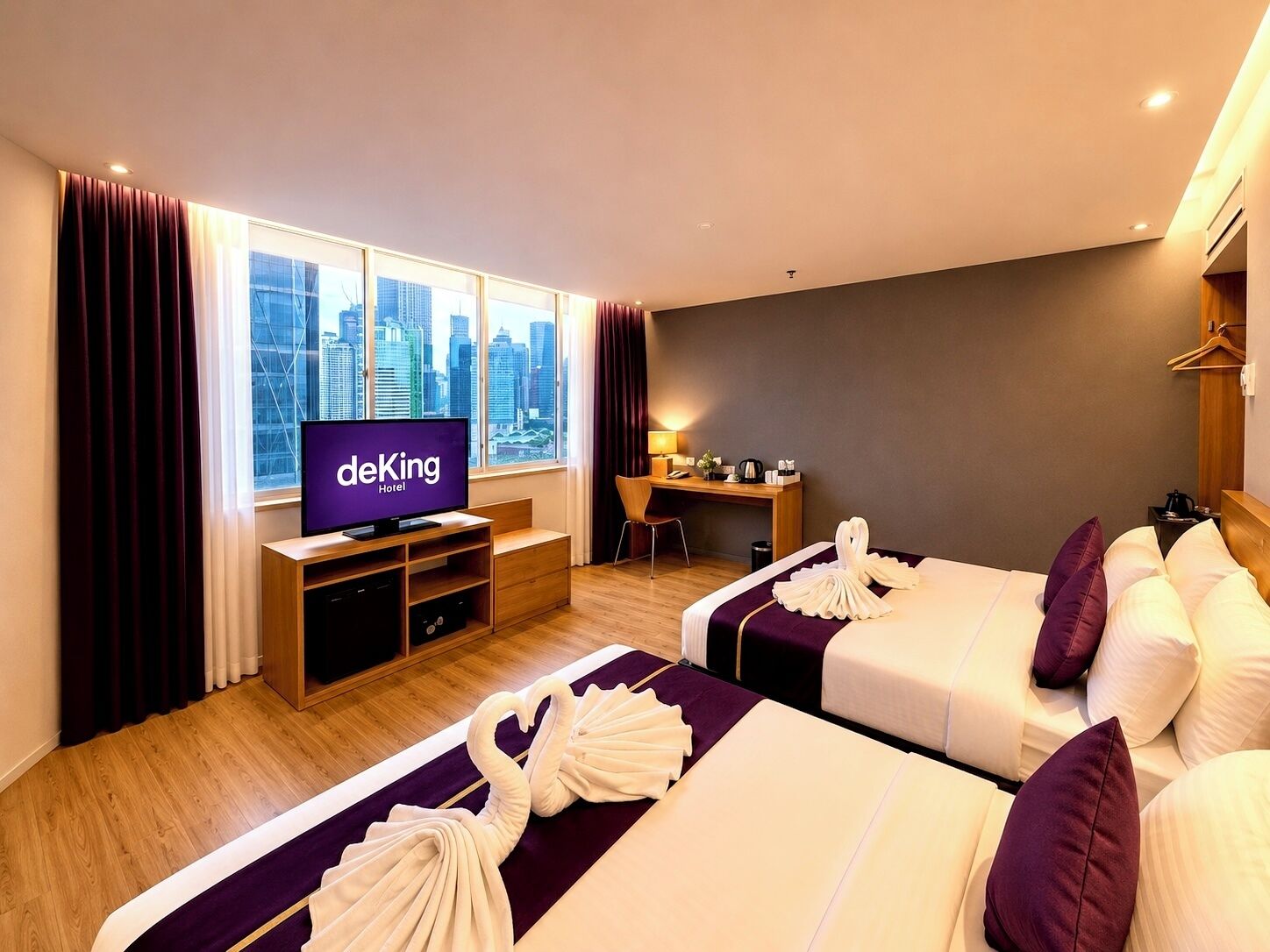 A hotel room with a television displaying 'deKing Hotel,' a desk with a chair, two beds with towels, and a city view.