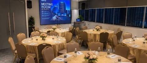 A meeting room with multiple round tables, chairs, and a large screen displaying a presentation.