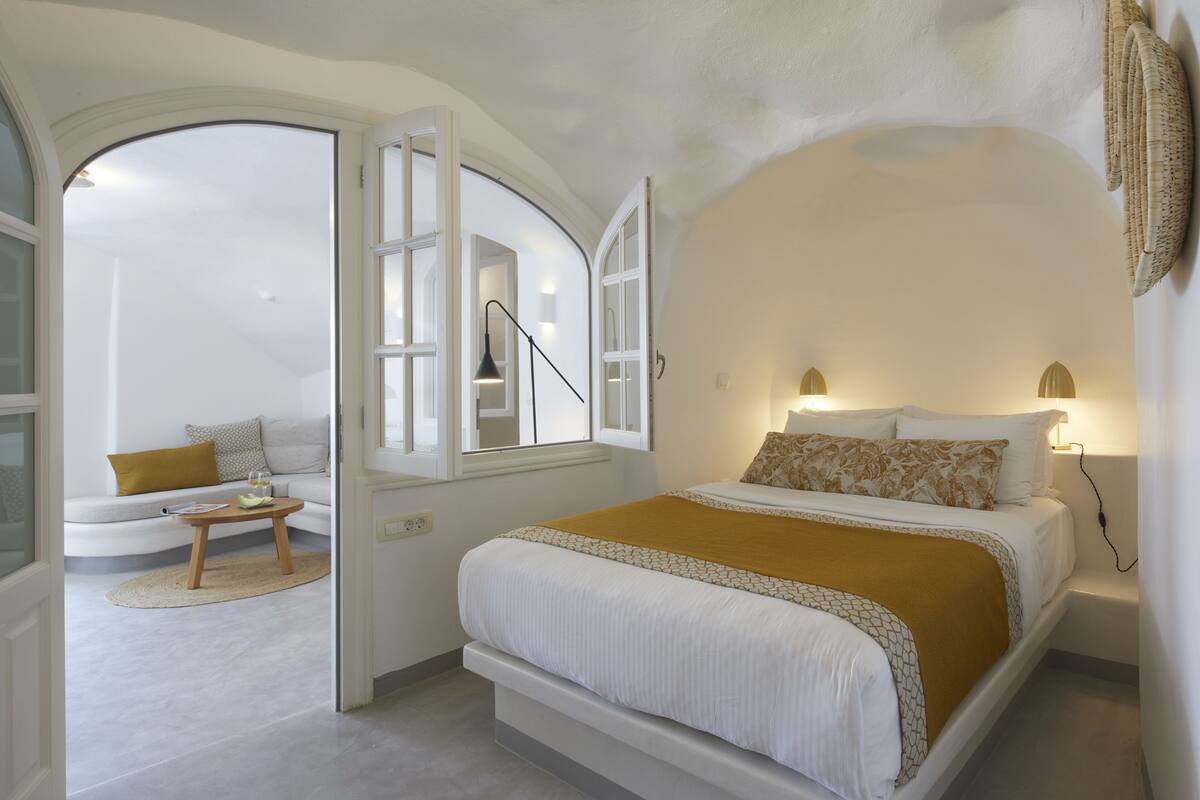 Un dormitorio con cama, lámparas de noche, una mesita de noche, una pequeña mesa con lámpara y vistas a una sala de estar con sofá y ventana.