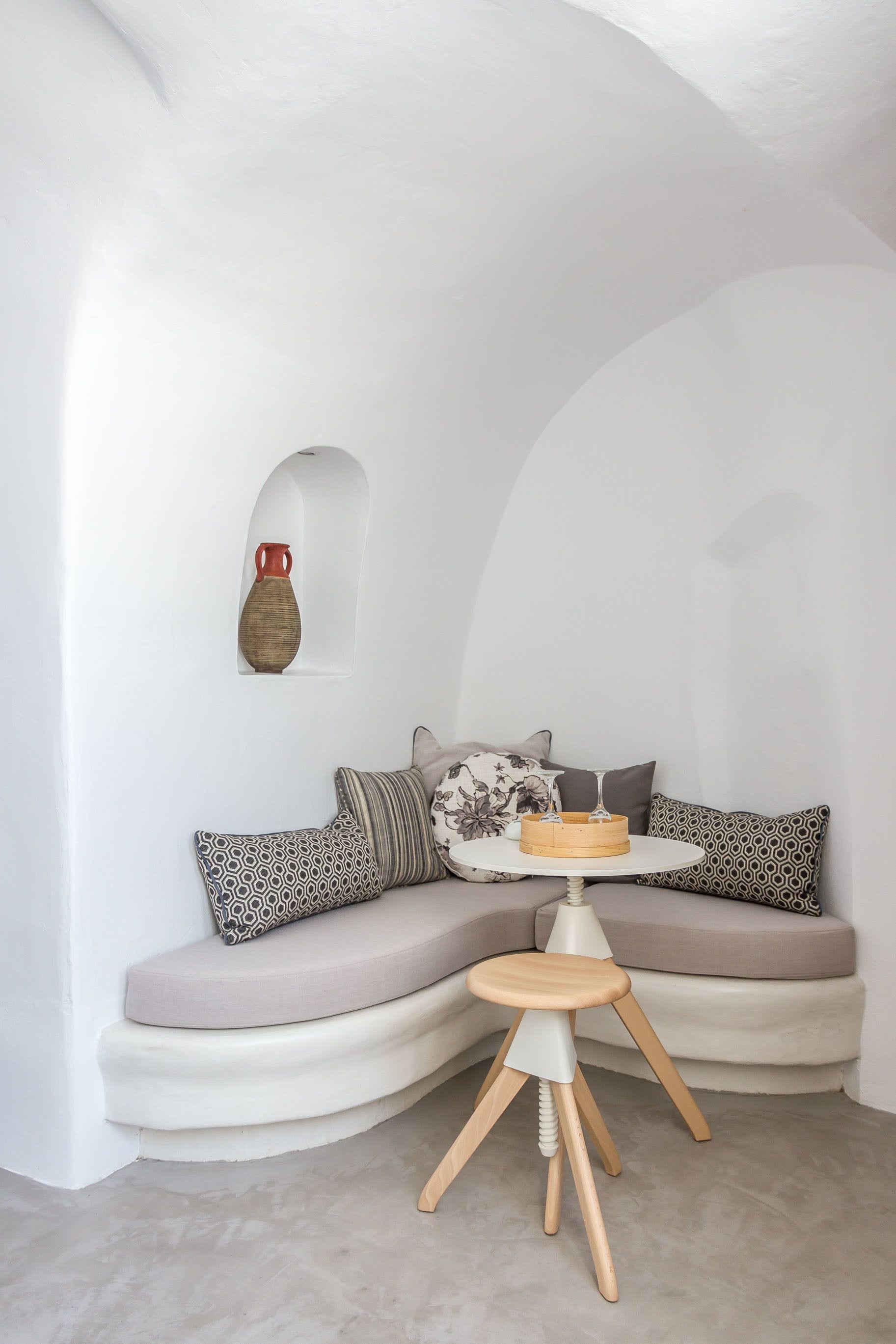 A cozy corner with a curved white sofa, a small wooden stool, and a vase on the wall.
