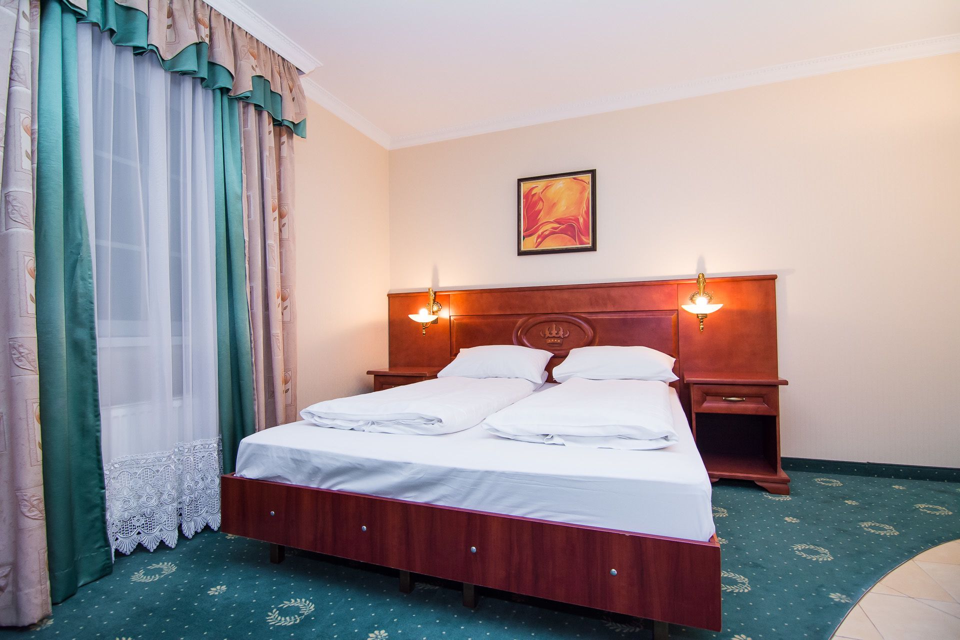 A hotel room with a wooden bed, two bedside tables, a nightstand, a painting on the wall, and a green carpet.