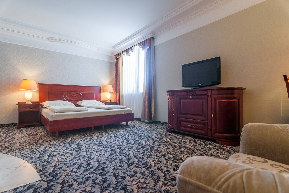 A hotel room with a bed, two bedside lamps, a television on a cabinet, and a patterned carpet.