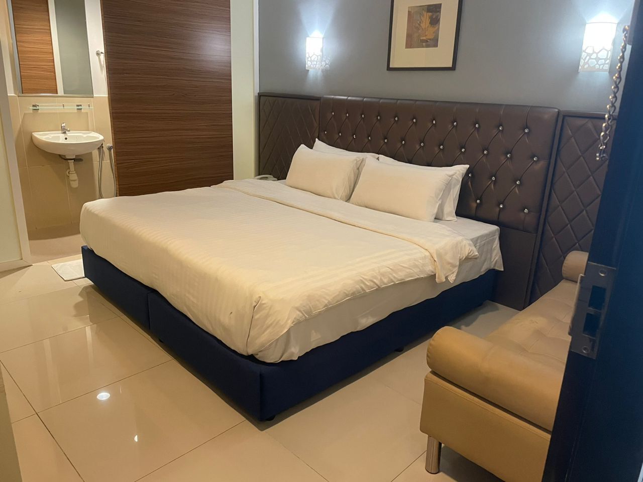 A bedroom with a large bed, a bedside table, a chair, a wall-mounted light, and a small sink.