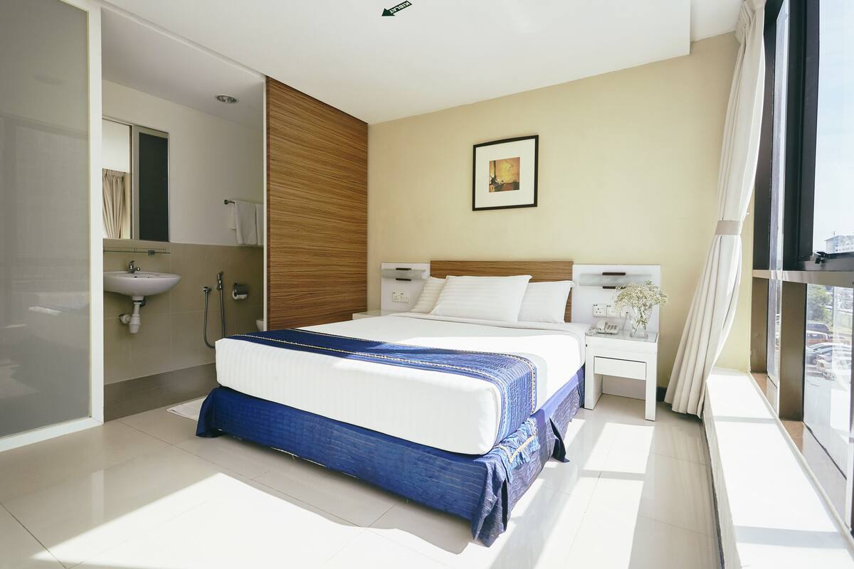Een moderne hotelkamer met een groot bed, een nachtkastje, een raam met gordijnen en een badkamer met een wastafel en spiegel.
